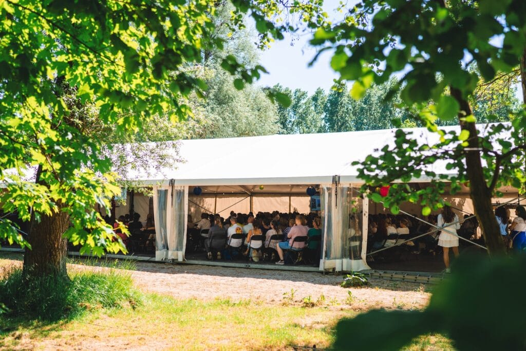 aluhal-congres-bos-groen-natuur Een bijzondere vergaderlocatie in de natuur met een aluhal waar mensen in moderne sfeer luisteren naar een spreker – even wat anders als een strand locatie, rolstoeltoegankelijk en perfect voor een congrescentrum met capaciteit op maat.